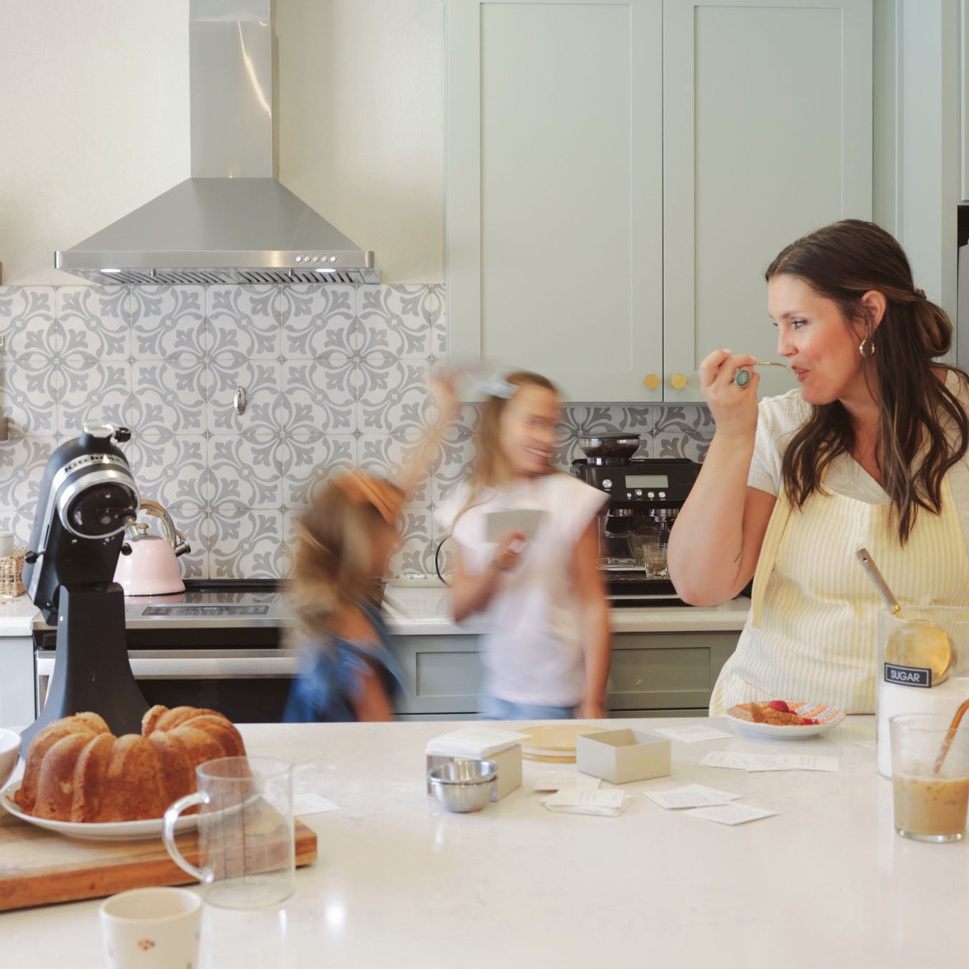 A mother and her two young daughters enjoy time together in a bright, modern kitchen. The mother is smiling and holding a spoon while preparing food, and the daughters move around the room. A bundt cake, coffee, and conversation cards are on the kitchen counter.