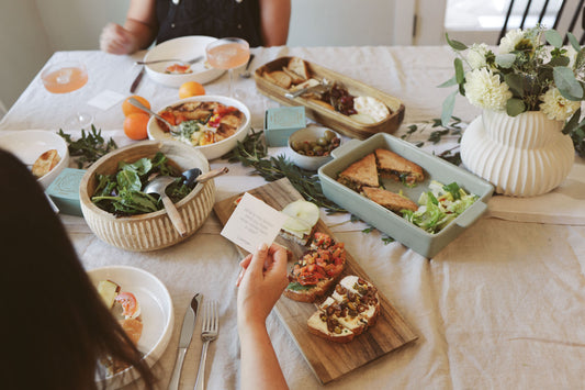 A beautifully set table with fresh, healthy dishes including sandwiches, salads, crostini, and olives, as well as glasses of a light pink drink. A person is holding a card from a set of Conversation Cards, sparking meaningful discussions among friends.