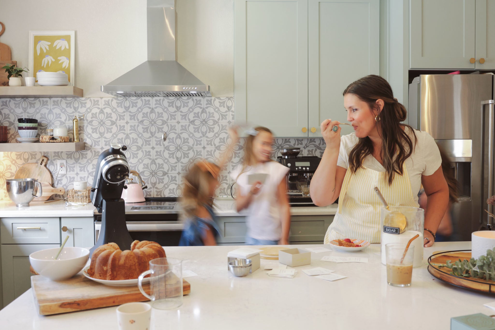 A mother and her two young daughters enjoy time together in a bright, modern kitchen. The mother is smiling and holding a spoon while preparing food, and the daughters move around the room. A bundt cake, coffee, and conversation cards are on the kitchen counter.