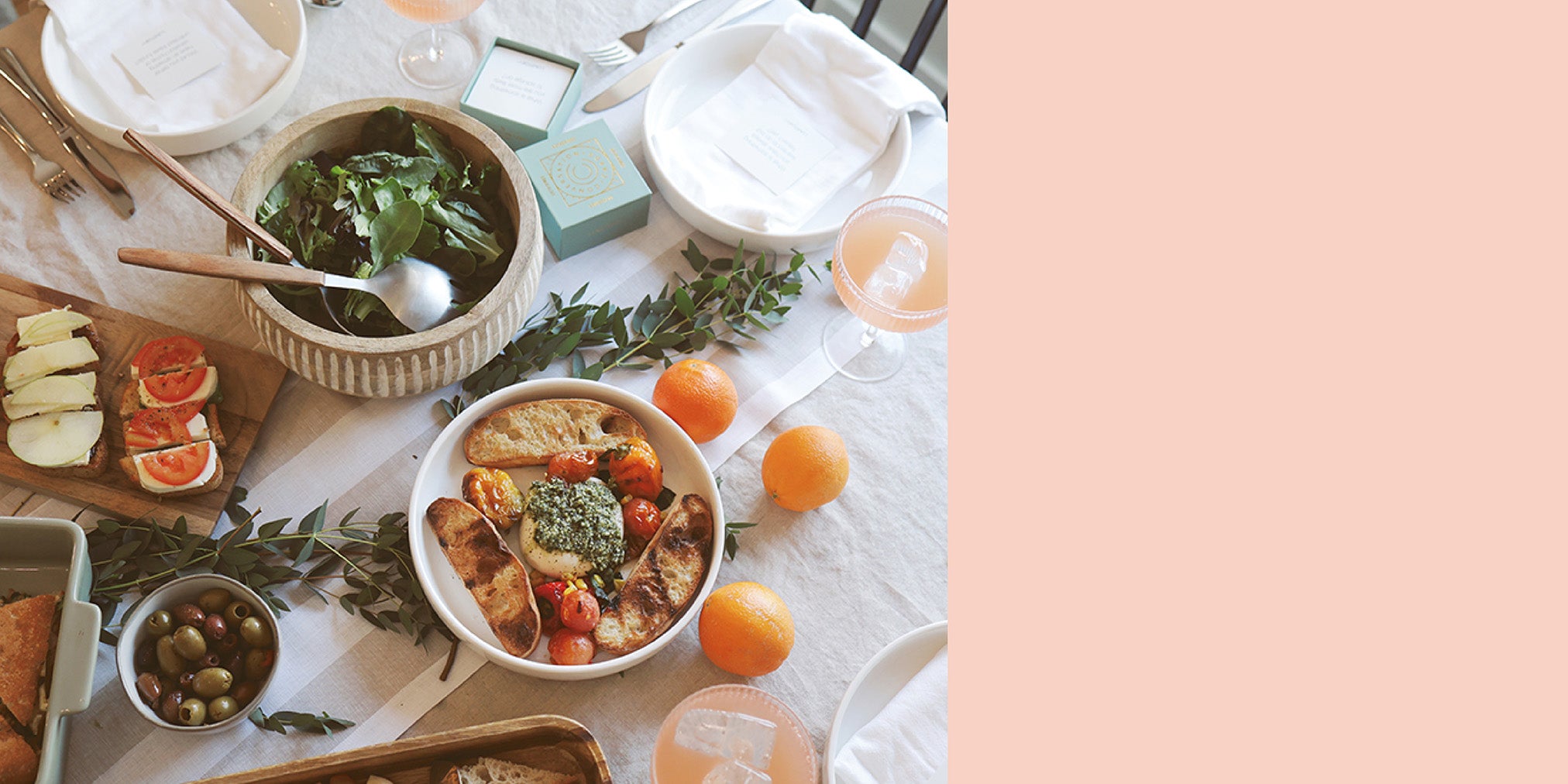 A beautifully arranged brunch table featuring a fresh salad, crostini with tomatoes and cheese, a bowl of olives, toasted bread with pesto, oranges, and a light cocktail, set on a neutral tablecloth with greenery for decoration.