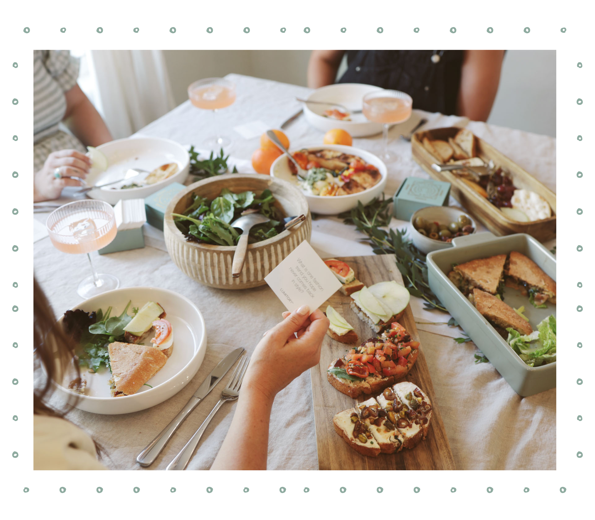 A beautifully set table with fresh, healthy dishes including sandwiches, salads, crostini, and olives, as well as glasses of a light pink drink. A person is holding a card from a set of Conversation Cards, sparking meaningful discussions among friends.